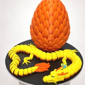 May include: A bright orange, scale-textured egg stands upright next to a long, articulated yellow dragon figure. The dragon has a segmented body, a detailed head with small blue eyes, and orange accents on its tail and head crest, resting on a circular base.