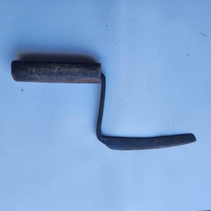 May include: A hand tool featuring a dark brown cylindrical wooden handle attached to a bent metal shank that connects to a flat, narrow, horizontal metal blade. The metal components appear weathered with a dark, matte, and slightly textured finish.
