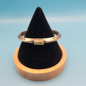 May include: A silver-toned metal ring featuring a rectangular, green and brown stone inlay at the center. The band is decorated with small, etched leaf-like patterns on either side of the stone, creating a detailed and textured appearance.