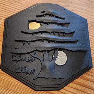 Könnte beinhalten: Schwarze, sechseckige Plakette mit einem Relief einer Zeder und zwei Münzen. Auf der Plakette befindet sich arabischer Text. Die Münzen sind gold- und silberfarben. Das Objekt liegt auf einer Holzoberfläche.