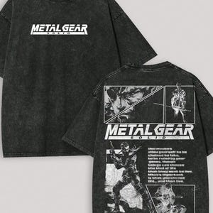 May include: A black acid-wash t-shirt featuring the white Metal Gear Solid logo on the chest. The back displays a large graphic print with multiple panels showing character illustrations, the Metal Gear Solid logo, and blocks of text.