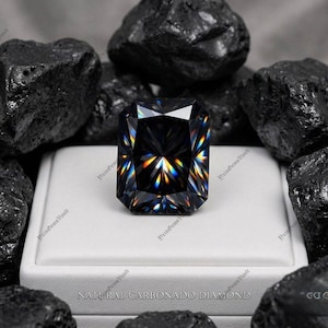 Peut inclure: Un diamant noir taille radiant avec une forte réfraction de la lumière affichant des éclats arc-en-ciel, posé sur une boîte de présentation blanche entourée de roches de carbone noir brut. Le texte "NATURAL CARBONADO DIAMOND" est imprimé.
