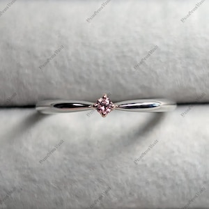 May include: A slender silver-toned ring featuring a single, small, round-cut pink gemstone set in a four-prong rose gold setting at the center. The band has a smooth, polished finish and tapers slightly toward the central stone.