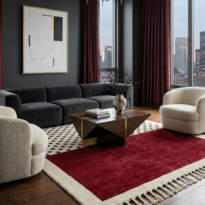 Puede incluir: Una sala de estar con un sofá gris oscuro, dos sillones texturizados de color crema y una mesa de centro geométrica de madera con detalles en latón. Una alfombra rectangular grande con centro rojo intenso y borde crema con flecos.