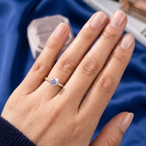 Puede incluir: Un anillo de oro con una piedra preciosa redonda de color violeta claro en el centro, con una banda acentuada por pequeñas piedras transparentes en los lados. El anillo se lleva en un dedo, mostrando la banda de oro metálico y la piedra brillante.