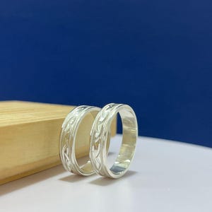 Puede incluir: Dos anillos de metal color plata con un patrón en relieve continuo de formas entrelazadas similares a hojas a lo largo de la banda exterior, bordeados por finos bordes de cuentas en relieve en la parte superior e inferior.