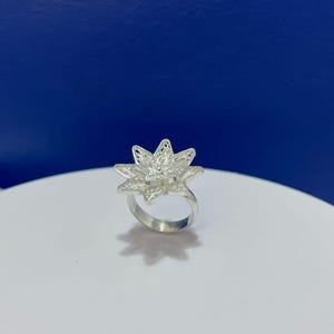 Puede incluir: Un anillo de plata con un diseño de flor grande de múltiples pétalos, elaborado con intrincado trabajo de filigrana. El metal tiene un acabado brillante y reflectante, y el centro floral se compone de pétalos puntiagudos superpuestos.