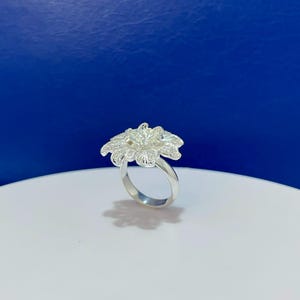 Puede incluir: Un anillo de tono plateado que presenta un gran diseño floral de varias capas. Los pétalos exhiben una textura de filigrana fina e intrincada en toda la superficie, creando una apariencia botánica detallada. La banda es lisa y pulida.