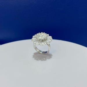 Puede incluir: Un anillo de plata con un diseño floral grande de varias capas. Los pétalos están elaborados con un intrincado trabajo de filigrana de hilo fino, creando una textura delicada y calada. El centro de la flor está elevado, añadiendo profundidad.