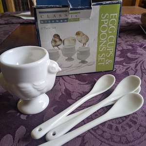 Könnte beinhalten: Ein weißer Eierbecher aus Keramik in Form eines Huhns, begleitet von drei weißen Keramiklöffeln. Die Artikel werden vor einem Pappkarton mit der Aufschrift Classic Kitchen 8 Piece Set Egg Cups & Spoons Set und Kükenbildern präsentiert.