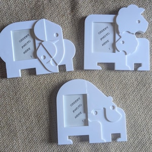 May include: Three white animal-shaped photo frames made of a smooth material. Each frame features a cutout for a picture with the text INSERT PHOTO HERE printed inside. The designs include an elephant, a lion with a cub, and a bear with a cub.
