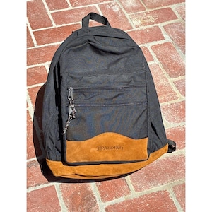 May include: A black backpack featuring a large main compartment and a smaller front pocket with two horizontal zippered openings. The base and a curved lower front panel are made of tan suede, with the Spalding logo embossed on the front panel.