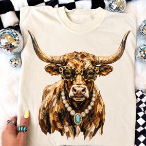 May include: A cream-colored t-shirt featuring a graphic print of a highland cow with long horns. The cow is wearing leopard print glasses and a necklace with a large, central turquoise-colored stone surrounded by smaller clear, sparkling gems.