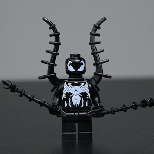 Può includere: Una minifigure in plastica nera con un emblema a ragno bianco sul busto e segni oculari bianchi sulla testa. La figura ha due grandi appendici nere curve e spinose sulla schiena e tiene in mano due lunghe aste nere sottili e segmentate.