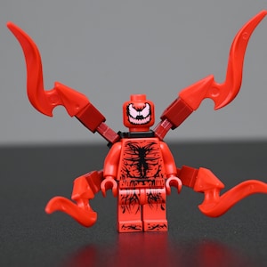 Puede incluir: Una minifigura de plástico roja con un patrón impreso en negro en el torso y las piernas, que presenta una cara sonriente en blanco y negro. La figura tiene cuatro apéndices rojos articulados en la espalda, cada uno con una punta curva.