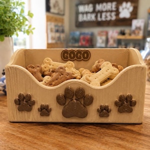 K&ouml;nnte beinhalten: Ein hellbeiger, rechteckiger Leckerlibeh&auml;lter aus Holz mit einem geschnitzten Pfotenabdruck auf der Vorderseite und dem eingepr&auml;gten Namen COGO oben in der Mitte. Der Beh&auml;lter ist mit knochenf&ouml;rmigen Hundeleckerlis in Hell- und Dunkelbraun gef&uuml;llt.