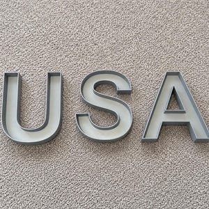 Könnte beinhalten: Drei einzelne dreidimensionale Buchstaben, die das Wort USA bilden. Jeder Buchstabe besteht aus einem soliden hellgrauen Material mit glatter Oberfläche und verfügt über ein tiefes, offenes Kanaldesign mit erhöhten Kanten.