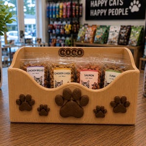 K&ouml;nnte beinhalten: Ein helles Holzregal mit der Aufschrift COCO und mehreren braunen Pfotenabdruck-Ausschnitten auf der Vorderseite. Darin befinden sich vier durchsichtige Plastikt&uuml;ten mit Katzenleckerlis, beschriftet mit H&uuml;hnchenbrust, H&uuml;hnerleber, Lachs und Thunfisch.