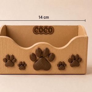 K&ouml;nnte beinhalten: Ein hellbrauner, rechteckiger Aufbewahrungsbeh&auml;lter mit Holzmaserung, verziert mit dunkelbraunen Pfotenabdr&uuml;cken auf der Vorderseite und dem eingepr&auml;gten Namen COCO an der Innenwand. Er ist 14 cm breit und 7 cm hoch.
