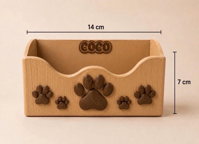 K&ouml;nnte beinhalten: Ein hellbrauner, rechteckiger Aufbewahrungsbeh&auml;lter mit Holzmaserung, verziert mit dunkelbraunen Pfotenabdr&uuml;cken auf der Vorderseite und dem eingepr&auml;gten Namen COCO an der Innenwand. Er ist 14 cm breit und 7 cm hoch.