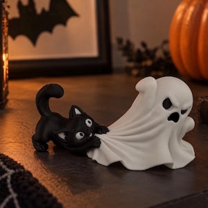 Könnte beinhalten: Eine dekorative Figur, die eine kleine schwarze Katze mit großen Augen zeigt, die spielerisch an dem weißen Laken eines Geistes zieht. Der Geist hat einen grimmigen Ausdruck mit dunklen, hohlen Augen und Mund, während die Katze geduckt ist.