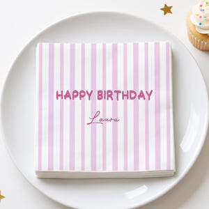 May include: A stack of square paper napkins featuring a light pink and white vertical striped pattern. The text "HAPPY BIRTHDAY" is printed in a bold pink font, followed by the name "Laura" in a cursive pink script font.