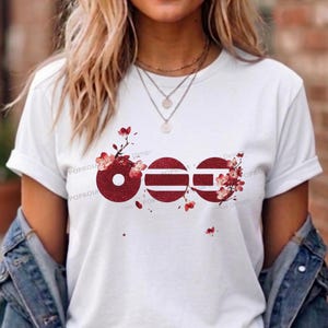 May include: A white short-sleeved t-shirt featuring a graphic design of three circular shapes in a deep red glitter texture. The circles are adorned with delicate pink and white cherry blossom branches and flowers, creating a floral motif.