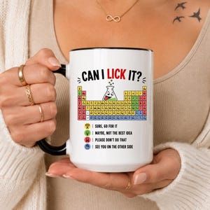 Peut inclure: Un mug en céramique blanc avec une anse et un bord noirs, orné d'un tableau périodique, d'une fiole de dessin animé et du texte CAN I LICK IT? accompagné d'une légende codée par couleur avec des icônes de trophée, ampoule, crâne et main.