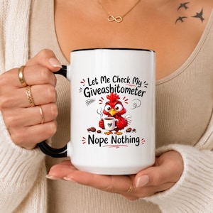 Pode incluir: Uma caneca de cerâmica branca com alça e borda pretas, apresentando um gráfico de um frango de desenho animado vermelho segurando uma pequena xícara de café. O texto diz: Let Me Check My Giveashitometer, Nope Nothing. Grãos de café espalhados cercam o frango.