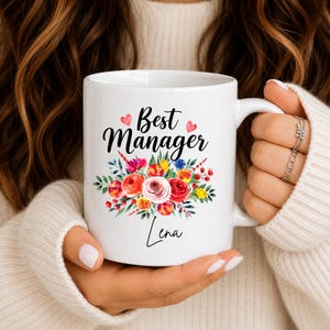 Puede incluir: Una taza de cerámica blanca con el texto en negro Best Manager y el nombre Lena en fuente cursiva. El diseño incluye un colorido arreglo floral en acuarela de flores rojas, rosas y amarillas con hojas verdes y dos pequeños corazones rosas.