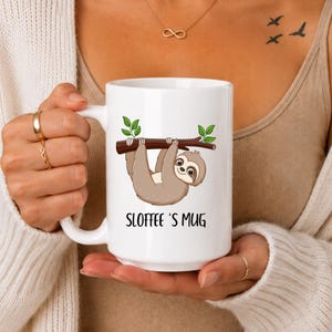 Peut inclure: Un mug en céramique blanche orné d'une illustration de paresseux brun suspendu à une branche d'arbre avec des feuilles vertes. Sous le paresseux, le texte SLOFFEE'S MUG est inscrit dans une police noire simple. Le mug a une finition lisse.