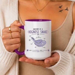 Puede incluir: Una taza de cerámica blanca con interior y asa de color morado, que presenta un gráfico titulado Anatomy of a Hognose Snake. El diseño incluye una ilustración de una serpiente morada con etiquetas que señalan partes del cuerpo.