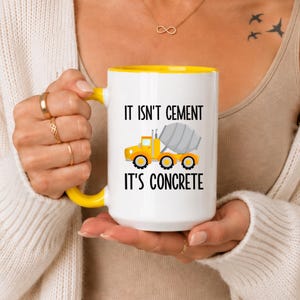 Peut inclure: Un mug en céramique blanche avec une anse et un bord intérieur jaune vif. L'avant présente un graphique d'un camion toupie jaune avec le texte noir "IT ISN'T CEMENT" au-dessus et "IT'S CONCRETE" imprimé en dessous du camion.