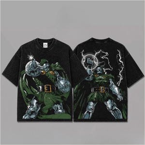Puede incluir: Una camiseta negra de manga corta con un estampado gráfico de un personaje enmascarado con capa verde y armadura metálica. El frente muestra al personaje señalando hacia adelante, mientras que la espalda muestra al personaje con el puño en alto.