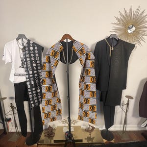 May include: Three garments displayed on hangers: a white t-shirt with black graphic text, a black vest with white Adinkra symbols, a central yellow and black patterned fabric stole with a gold chain, and a black blazer with a textured lapel.