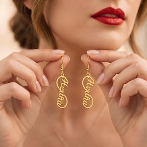 May include: A pair of gold-toned drop earrings featuring a vertical infinity symbol design. Each earring displays the name Hertha in a cursive script font integrated into the center of the infinity loop, suspended from a simple gold hook wire.
