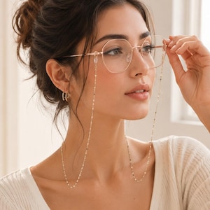 Op de afbeelding: Goudkleurige metalen bril met een geometrische zeshoekige montuurvorm. De bril is bevestigd aan een fijne, platte gouden schakelketting die vanaf de veren naar beneden hangt, wat een functioneel en decoratief accessoire voor de bril vormt.