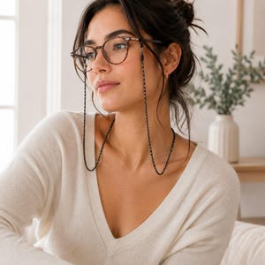 Puede incluir: Unas gafas de carey marrón con una cadena negra fina y texturizada sujeta a las patillas. La cadena cuelga en un bucle largo, con un patrón fino trenzado o de eslabones que descansa sobre la zona del pecho.