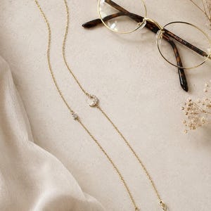 Op de afbeelding: Een brilketting van goudkleurig metaal met een fijn schakelontwerp. De ketting is versierd met een kleine, met heldere kristallen ingelegde maansikkel en een bijpassende sterbedel. Het heeft doorzichtige siliconen lussen aan beide uiteinden.