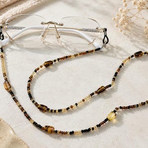 Op de afbeelding: Een bril zonder montuur met witte pootjes bevestigd aan een kralen brilketting. De ketting heeft een herhalend patroon van kleine zwarte, doorzichtige en amberkleurige kraaltjes, afgewisseld met grotere, langwerpige, doorschijnende amberkleurige kralen.