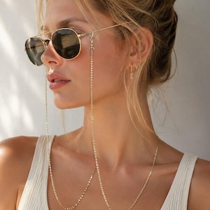 May include: A gold-toned metal eyeglass chain featuring a delicate link design, attached to the temples of dark-tinted aviator sunglasses. The chain hangs down in a long, slender loop, providing a functional and decorative accessory for eyewear.