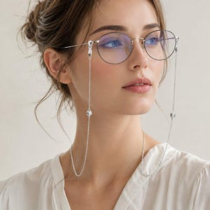 Peut inclure: Lunettes en métal argenté dotées de montures fines et rondes avec des verres transparents. Des chaînes argentées délicates sont fixées aux branches et tombent vers le bas, chacune ornée d'une petite perle en cristal transparent à facettes.
