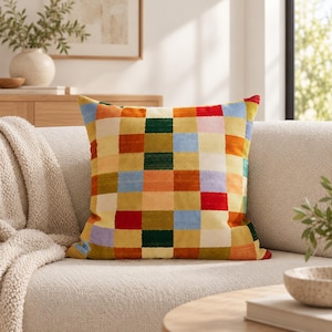 May include: A square throw pillow featuring a multi-colored checkered pattern. The design consists of small, uniform squares in shades of red, orange, yellow, green, blue, and cream, arranged in a grid across the entire surface of the pillow.