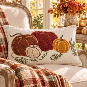 May include: A rectangular throw pillow with an off-white fabric base featuring a textured punch needle design of four pumpkins in shades of rust, deep red, cream, and golden yellow, accented with embroidered green vines and a stitched border.