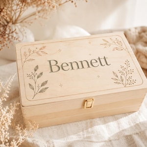 May include: A rectangular light-coloured wooden keepsake box featuring a hinged lid with a gold-tone metal latch. The lid is engraved with the name Bennett in the centre, surrounded by delicate botanical leaf illustrations and small star accents.
