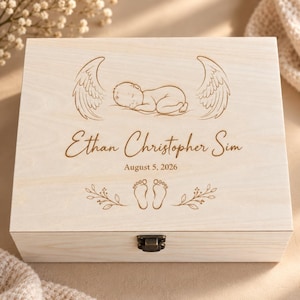 May include: A rectangular light-coloured wooden keepsake box with a metal latch. The lid features a laser-engraved design of a sleeping baby between two angel wings, the name Ethan Christopher Sim, the date August 5, 2026, and baby footprints.