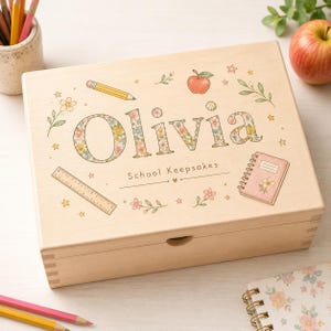 Può includere: Una scatola rettangolare in legno chiaro con il nome Olivia in lettere a motivi floreali. Il coperchio è decorato con illustrazioni di una matita, una mela, un righello, un taccuino e piccoli fiori. Il testo School Keepsakes è stampato sotto il nome.