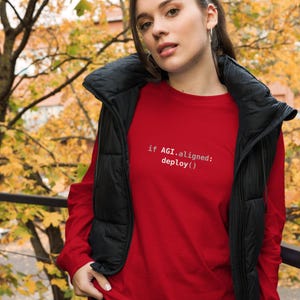 May include: A long-sleeve red shirt featuring the white text "if AGI.aligned: deploy()" printed on the chest, worn underneath a black sleeveless puffer vest with a high collar and a front zipper closure.