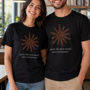 May include: A black short-sleeved t-shirt featuring a graphic of a brown sun-like shape composed of small dots. Below the graphic, the text reads "guess the next word" in a larger font, followed by "repeat until impressive" in a smaller font.