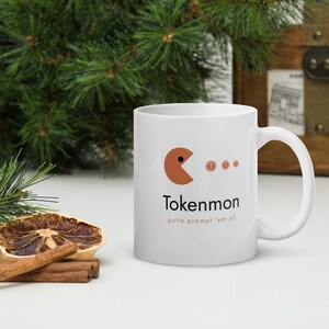 May include: A white ceramic mug featuring a brown circular character with a wedge-shaped mouth facing three small brown circular tokens. The text Tokenmon is printed in black below the character, with the phrase gotta prompt 'em all in smaller orange text.